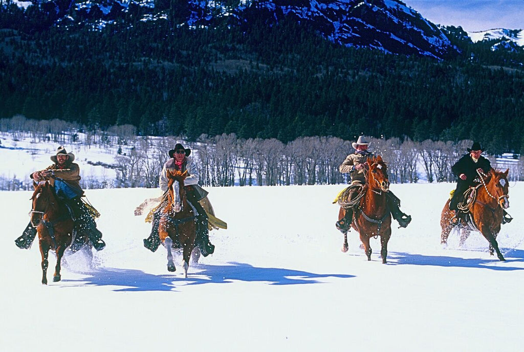 Four cowboys galloping through a snowy field with a majestic mountain range in the background.