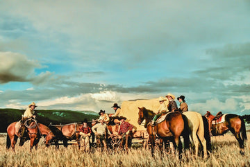 Art print of cowboys and cowgirls around a chuckwagon in the open plains with mountains and blue skies in the background.