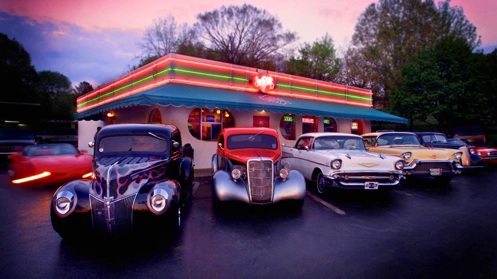 Classic car show at Mel's Diner during dusk, featuring a variety of vintage vehicles under glowing lights.