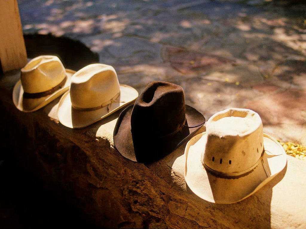 A collection of four cowboy hats on a rustic porch, each showcasing its unique crown, in a limited edition print by Michael W. Rutherford.