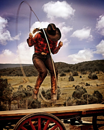 Cowgirl in Capitan, NM, trick-roping on a wagon, wearing a black hat and red plaid shirt against a backdrop of plains and hills.