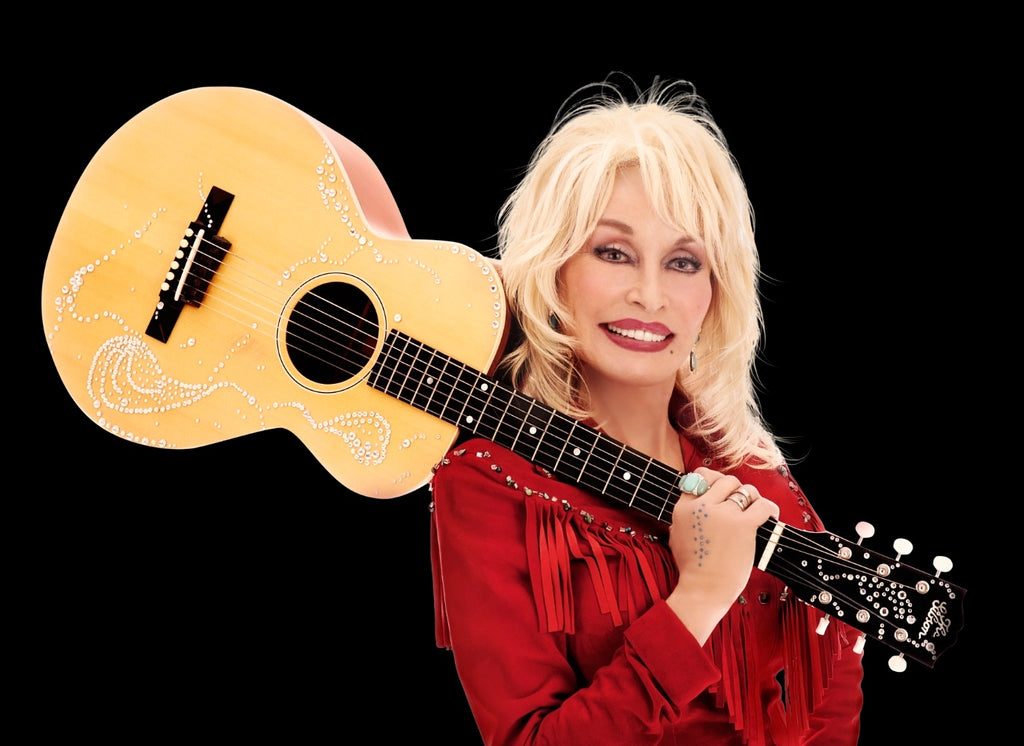 Dolly Parton in a red blouse, holding a guitar on her right shoulder with a black background.