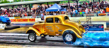 Vintage gold dragster at Beech Bend Raceway engulfed in blue smoke with a bustling crowd of spectators in the background.