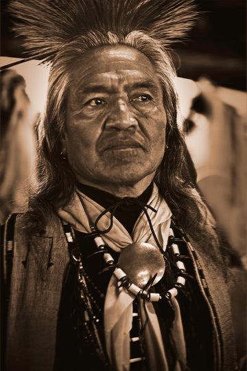 A sepia-toned portrait of an older Native American man. His expression serious and contemplative, face is lined with age, gray hair in a traditional style.