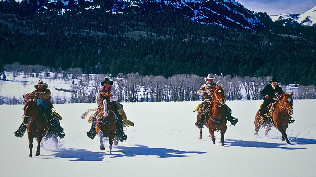 Four cowboys race their horses through deep snow. The snow-covered ground is level & the horses kick up snow in their wake.
