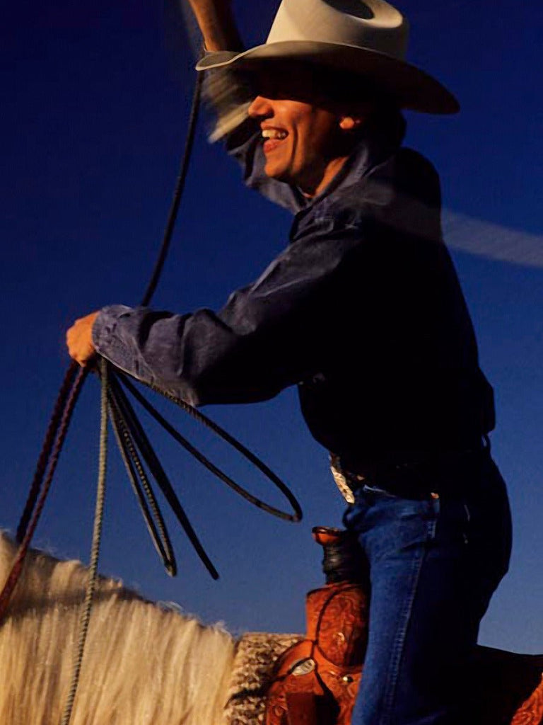 George Strait with lariat trick-roping on horseback at his Texas ranch, wearing blue jeans, a shirt, and a signature cowboy hat.