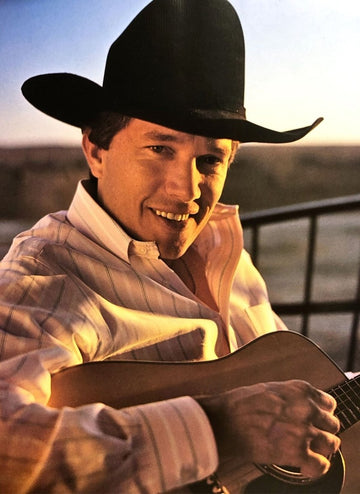 George Strait holding an acoustic guitar at his Texas ranch, wearing a white cowboy hat.