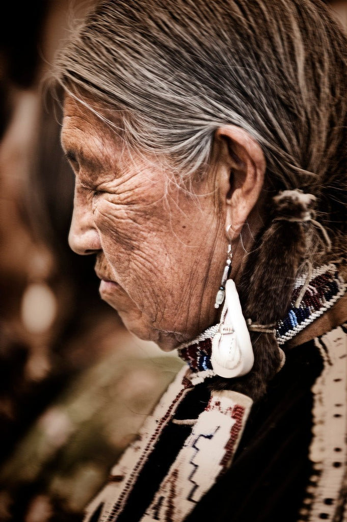 Elderly Native American woman in profile, weathered face is turned slightly downward, gray hair pulled back revealing a beaded choker around her neck.