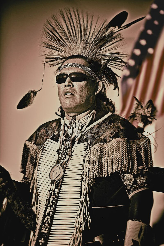 A Native American in mohawk-style regalia, sunglasses, with the sun shining and the American flag blurred in the background.