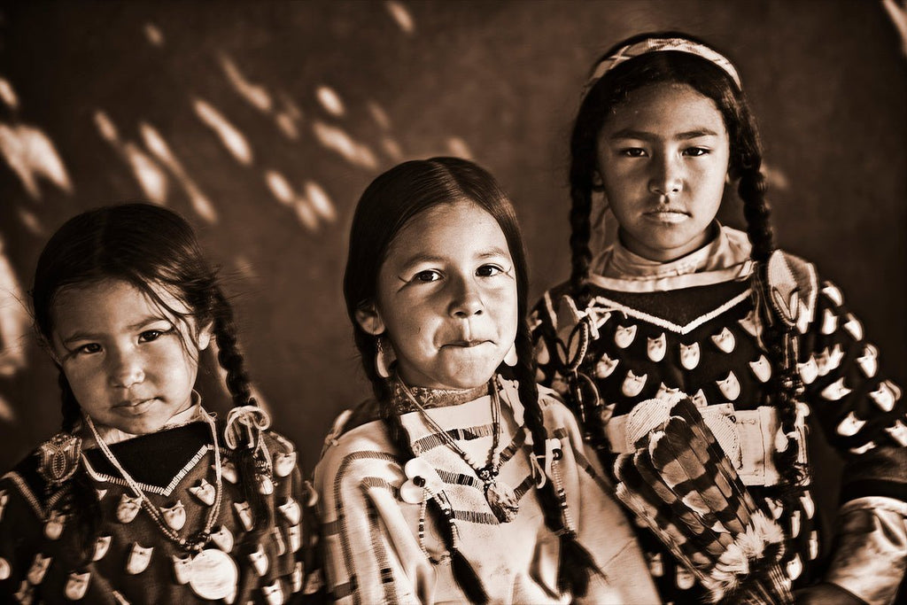 Three young Native American girls dressed in traditional clothing posed against a textured background in a vignette shot, wearing dresses with distinct patterns.