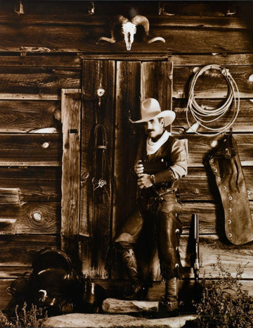 Sepia photo of cowboy with camera leaning on cabin with ram's skull door