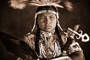 The image shows a young, likely adolescent, Native American individual dressed in traditional regalia, staring directly at the camera with a serious expression.