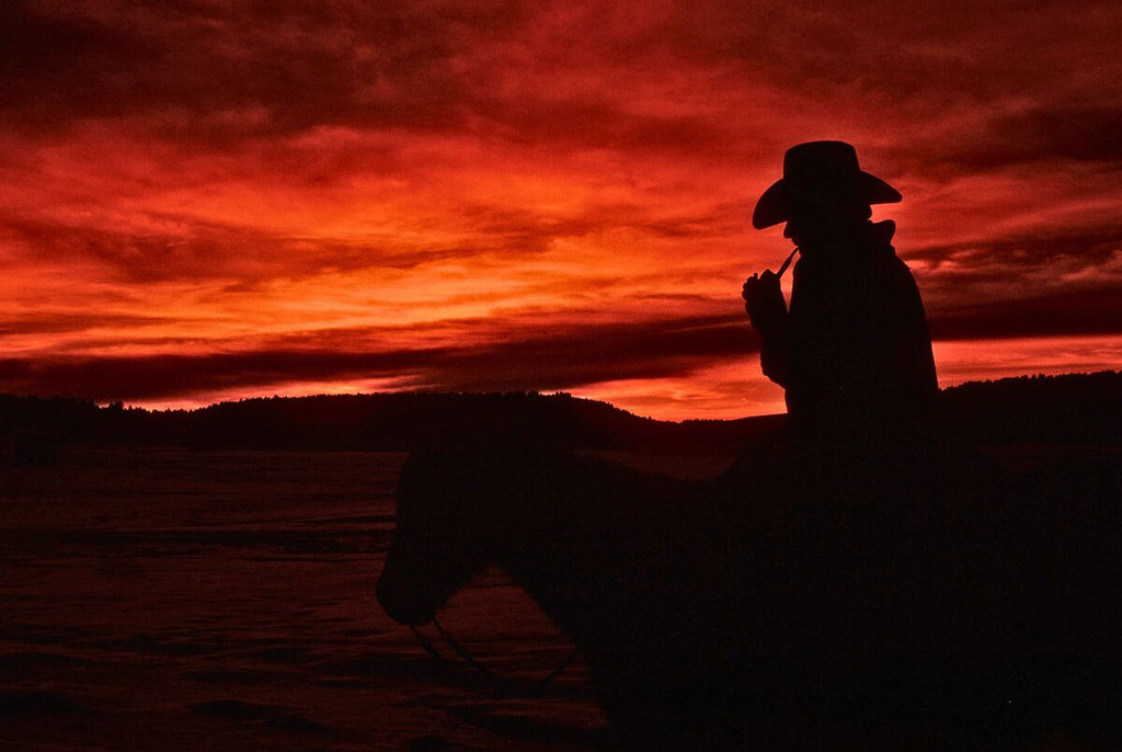 Silhouette of cowboy on horseback against a fiery red sunset with vast sky