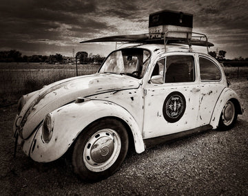 Vintage Volkswagen Beetle with a surfboard and storage container against an overcast sky in black and white image. 