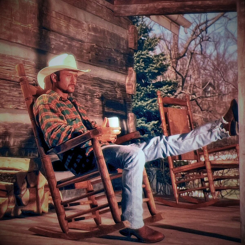 Tim McGraw sitting in rocking chair on a wooden porch of a log cabin, one foot propped up on a post, holding a mug.