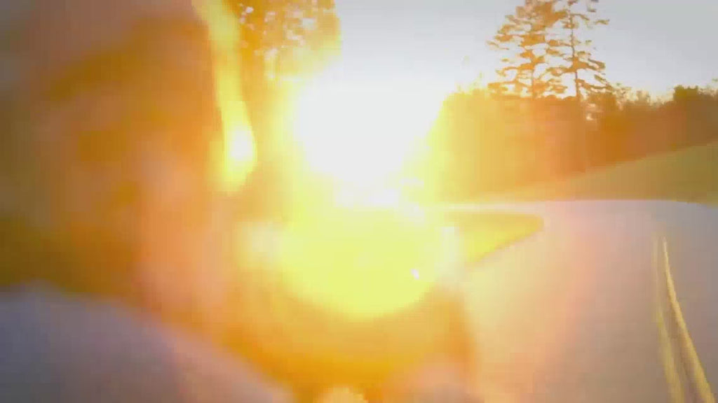 Video with sun shining on Rutherford holding camera in bed of truck, turning to show camera, motorcycle rider in background.